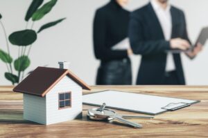 Model domu, klucze i dokumenty na stole, z rozmytymi postaciami w tle, symbolizujące zakup nieruchomości od syndyka.